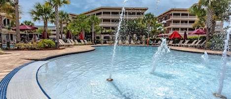 A swimming pool with a fountain, surrounded by palm trees and lounge chairs.