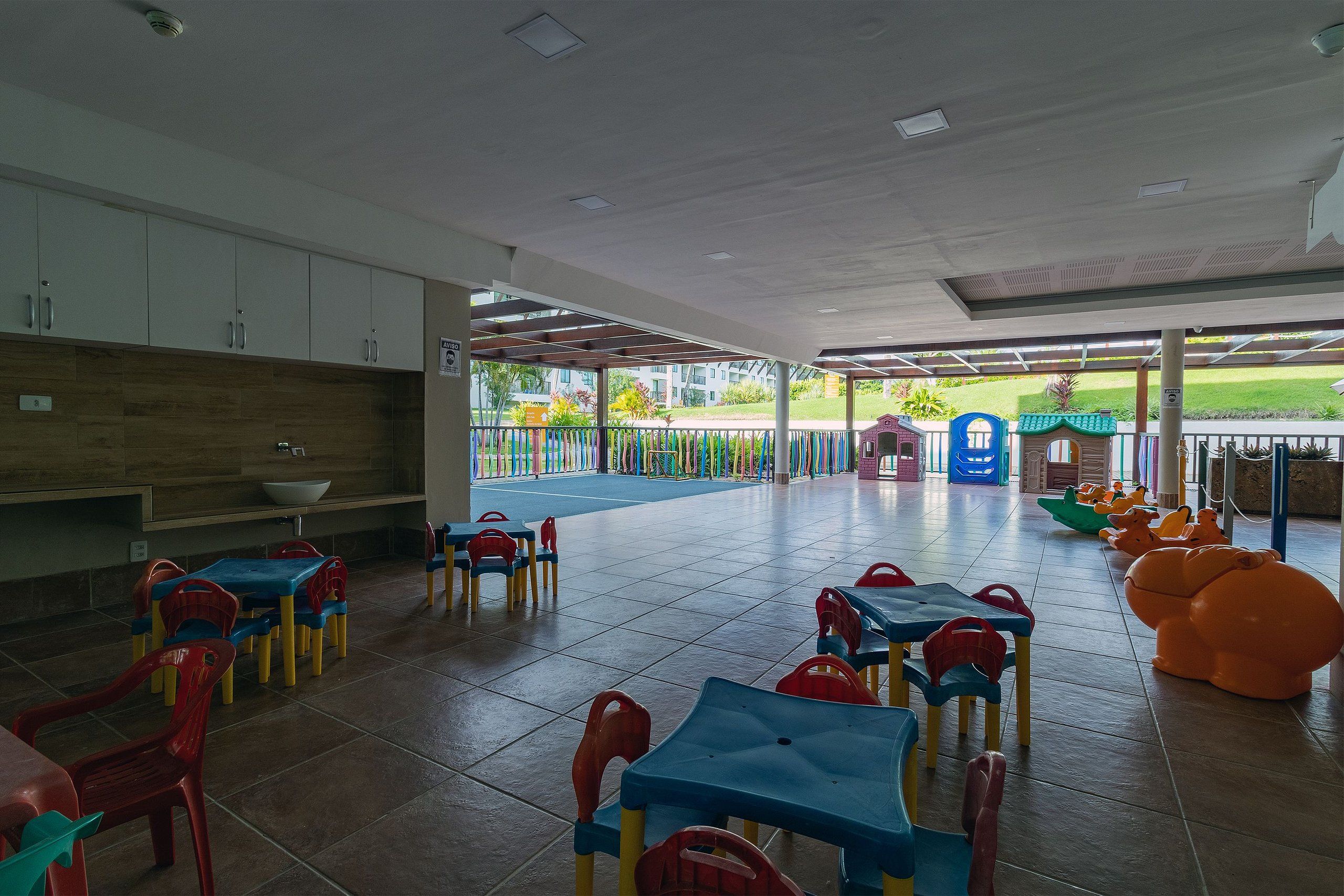 Zona de juegos infantiles con mesas y sillas de colores, un espacio de descanso cubierto y vistas al exterior.