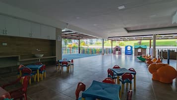 Zona de juegos infantiles con mesas y sillas de colores, un espacio de descanso cubierto y vistas al exterior.