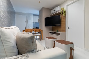 Una sala moderna con sofá, televisor de pantalla plana montado en la pared, comedor con mesa y sillas, y una cocina visible a través de un umbral abierto.
