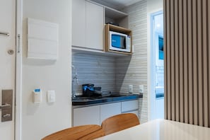 Cocina compacta con microondas, estufa y lavamanos. Hay una silla de madera y una mesa con mantel blanco.