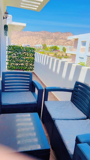 Eine Terrasse mit zwei Stühlen und einem Tisch, eine weiße Wand, ein grüner Heckenstreifen und Blick auf die Berge.
