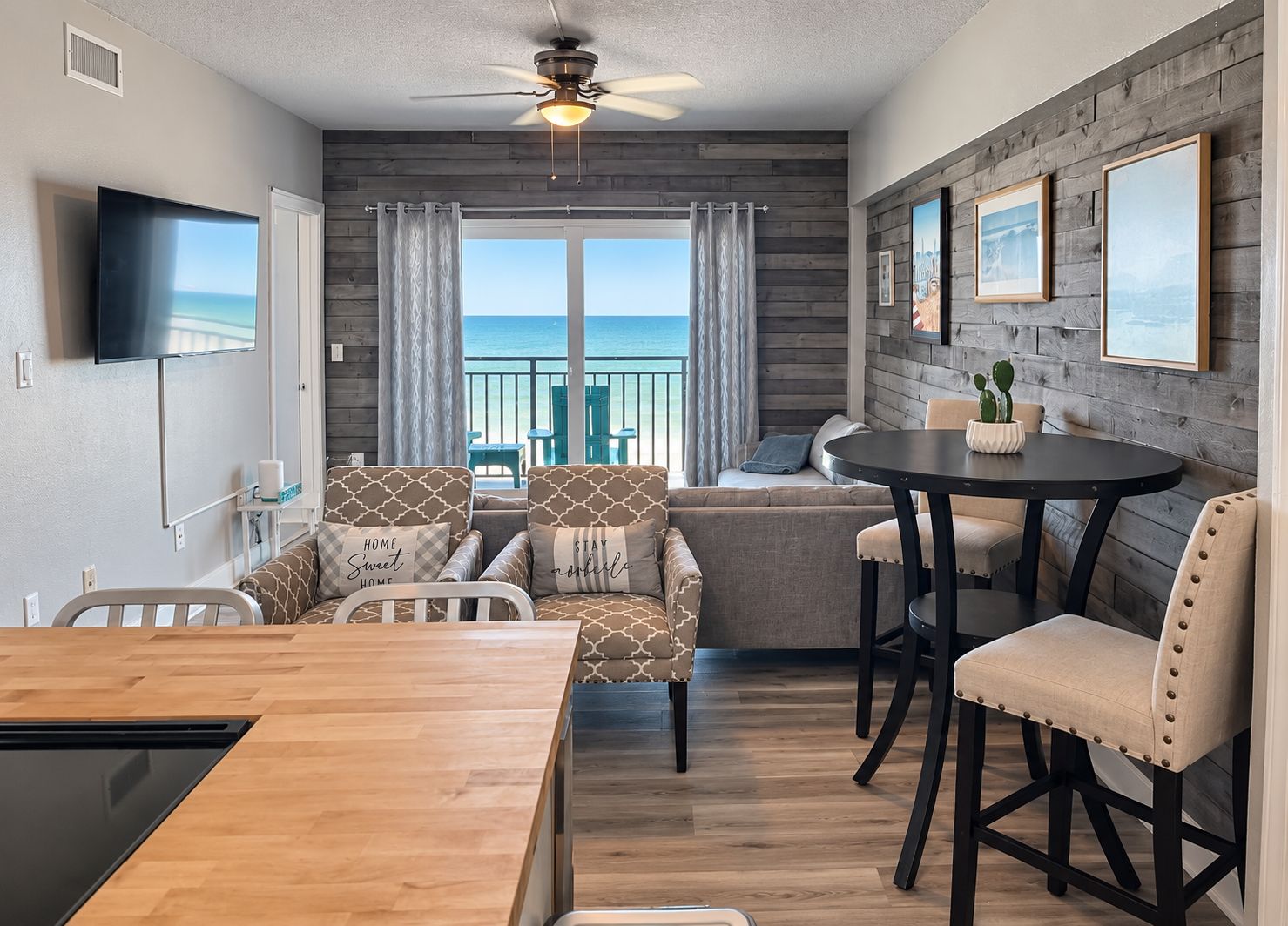 A room with a wooden ceiling fan, a television mounted on the wall, a dining table with chairs, a sofa with patterned cushions, and a view of the ocean through large windows.