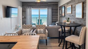 A room with a wooden ceiling fan, a television mounted on the wall, a dining table with chairs, a sofa with patterned cushions, and a view of the ocean through large windows.