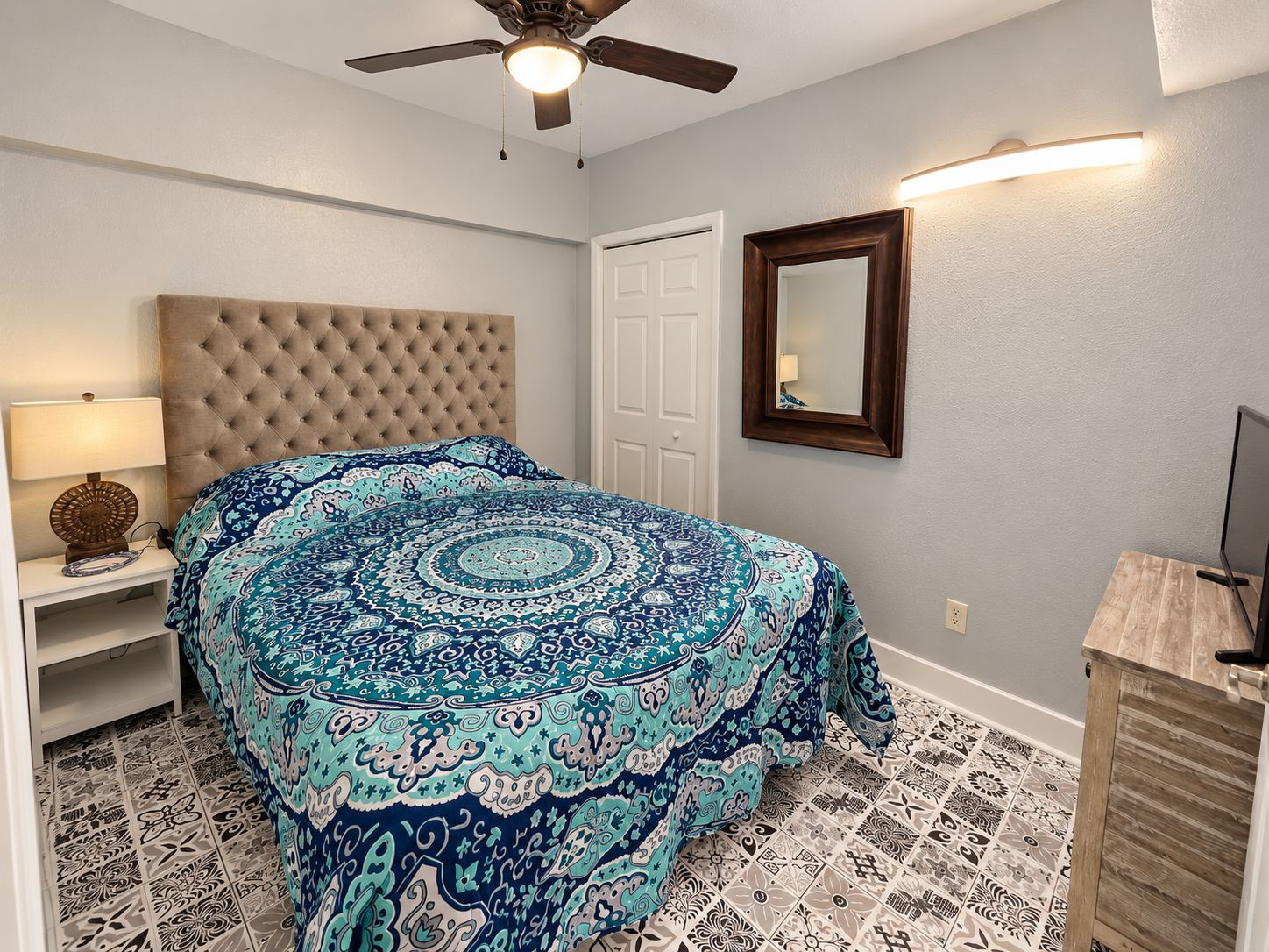 A bedroom with a tufted headboard, patterned bedspread, a bedside lamp, a ceiling fan, a mirror, and a wooden cabinet.