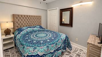 A bedroom with a tufted headboard, patterned bedspread, a bedside lamp, a ceiling fan, a mirror, and a wooden cabinet.