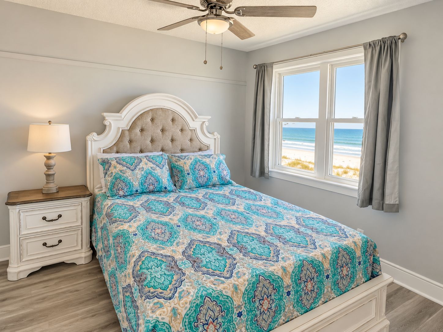 A bedroom with a bed, nightstand, lamp, and a view of the beach.