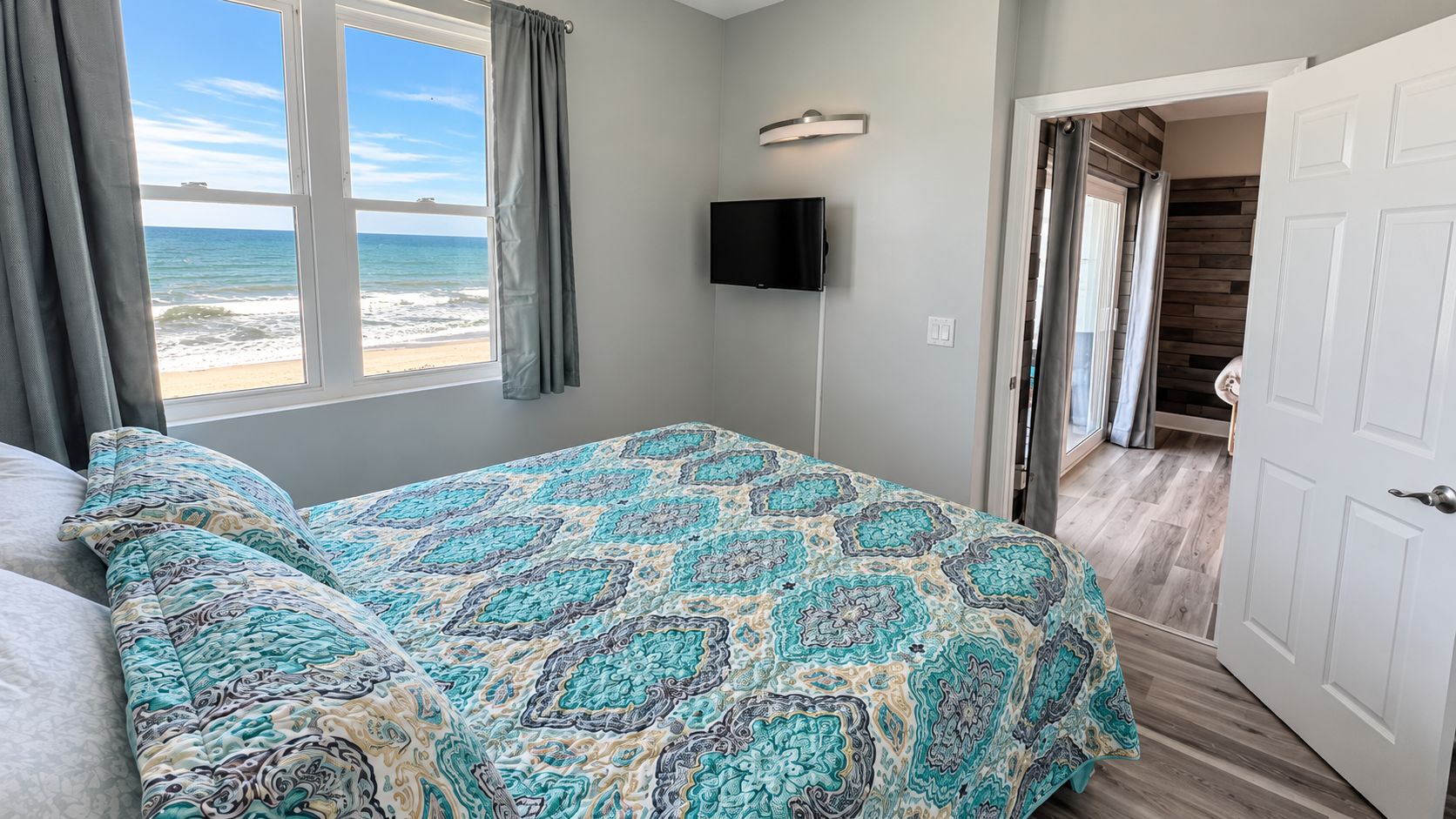 A bedroom with a bed, a TV, a view of the beach, and a door leading to a balcony.