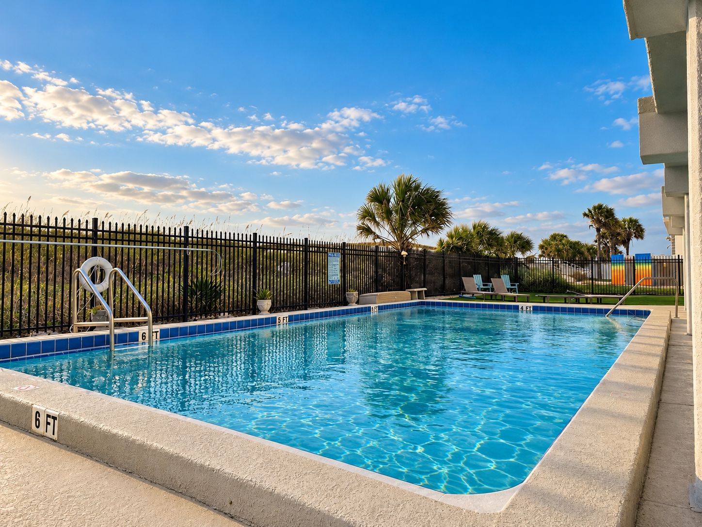 Une piscine rectangulaire entourée d’une clôture noire, avec des palmiers et un ciel dégagé.