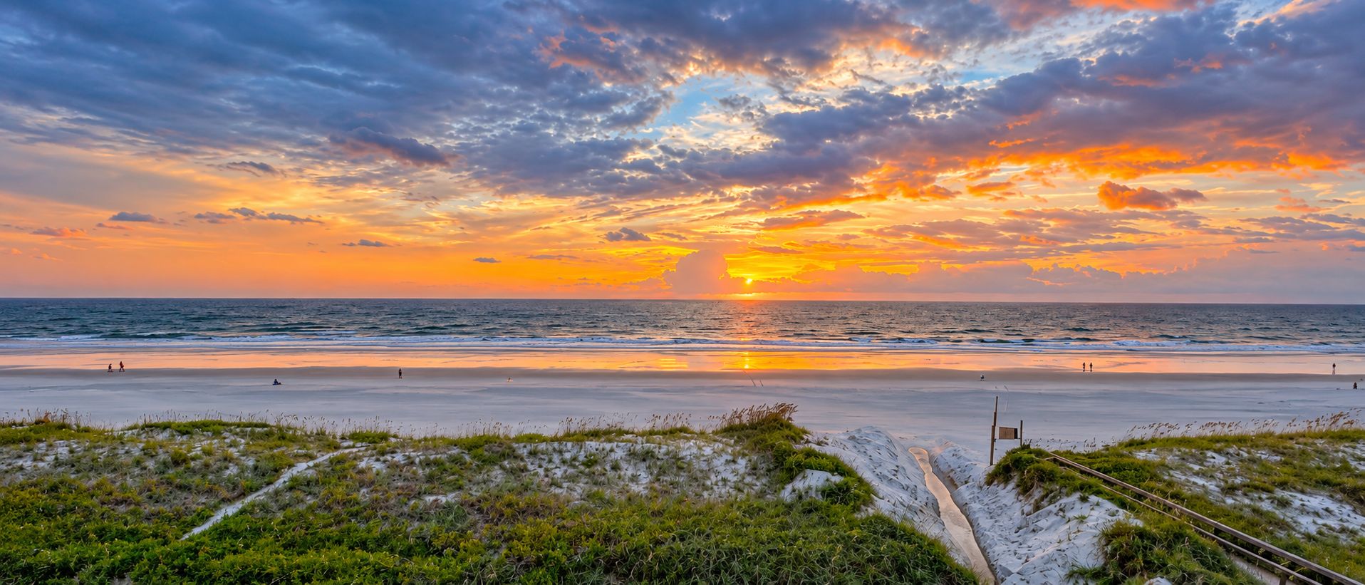 A beach with a sunset, waves, and a grassy dune area.