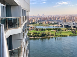 A high-rise building with a glass balcony overlooking a river, cityscape, and pyramids in the distance. - Vesta - Superior Suite - 2BR - Nile View (Cairo)