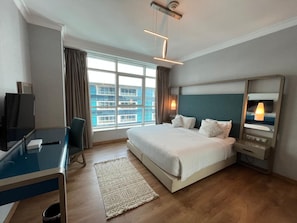 A hotel room with a large bed, a desk with a computer, a television, and a view of the city through the window. - Vesta - Superior Suite - 2BR - Nile View (Cairo)