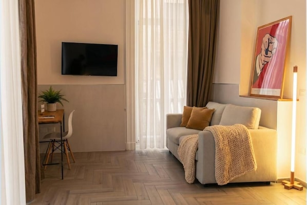 A modern living room with a sofa, a flat-screen TV, a desk with a chair, and a lamp. - Palazzo Bevilacqua Rooms (Naples)
