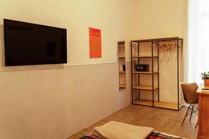 A room with a flat-screen TV mounted on the wall, a poster, a wooden floor, a foldable wardrobe, and a chair. - Palazzo Bevilacqua Rooms (Naples)