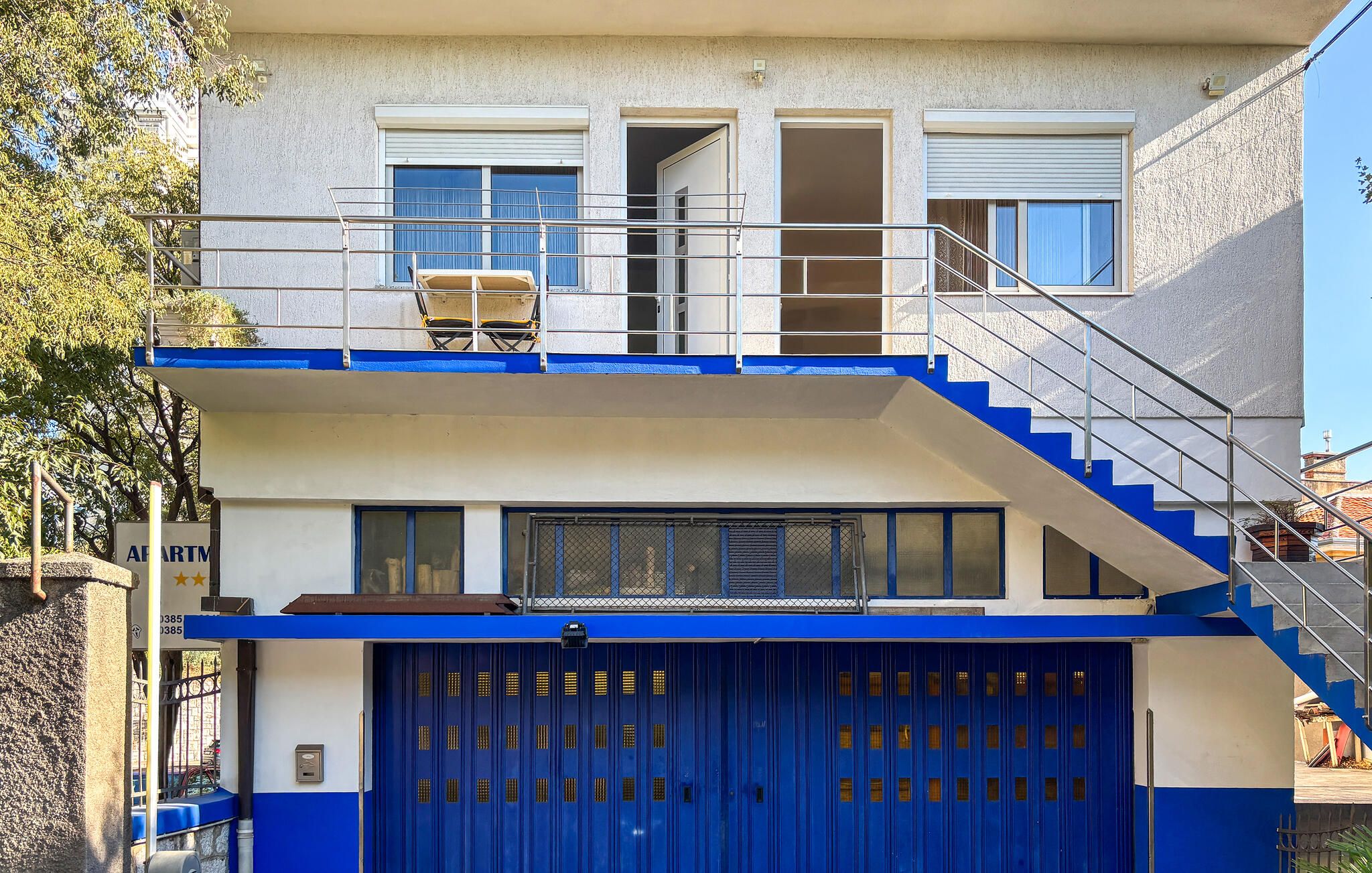 Un bâtiment à deux étages avec une porte de garage bleue, un balcon doté d’une rambarde en métal et un escalier menant à l’étage supérieur.