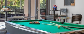 Une table de billard recouverte d’un drap vert, entourée de chaises et de tables, avec une vue sur l’extérieur par une fenêtre.