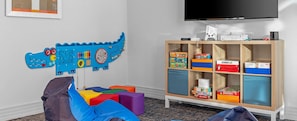 Un espace de jeux pour enfants avec un tapis de jeu bleu représentant un dinosaure, des blocs de mousse colorés et une étagère en bois contenant divers jeux.