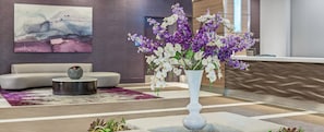 Eine moderne Lobby mit einer weißen Vase voller lila und weißer Blumen, einem grauen Sofa und einem gemusterten Teppich.