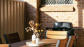 A wooden table with a white vase containing dried flowers, a grill, and a brick wall in the background. - Illa Villa is a charming townhouse just 50 meteres from Reddall Reserve. (Lake Illawarra)