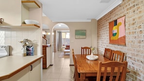A kitchen with a wooden table and chairs, a tiled backsplash, and a brick wall with a colorful painting. - Illa Villa is a charming townhouse just 50 meteres from Reddall Reserve. (Lake Illawarra)