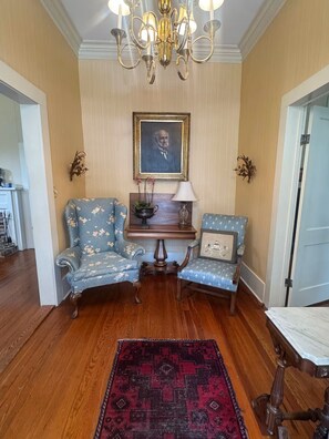 Une pièce comprenant deux fauteuils, une petite table avec une lampe, un portrait encadré et un tapis rouge à motifs.