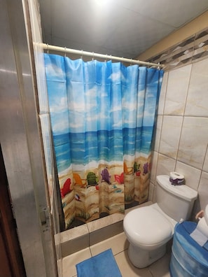 A bathroom with a shower curtain depicting a beach scene, a toilet, and a blue mat on the floor. - Affordable, self contained studio, 8-10 mins from the Piarco Intl Airport (Arima)