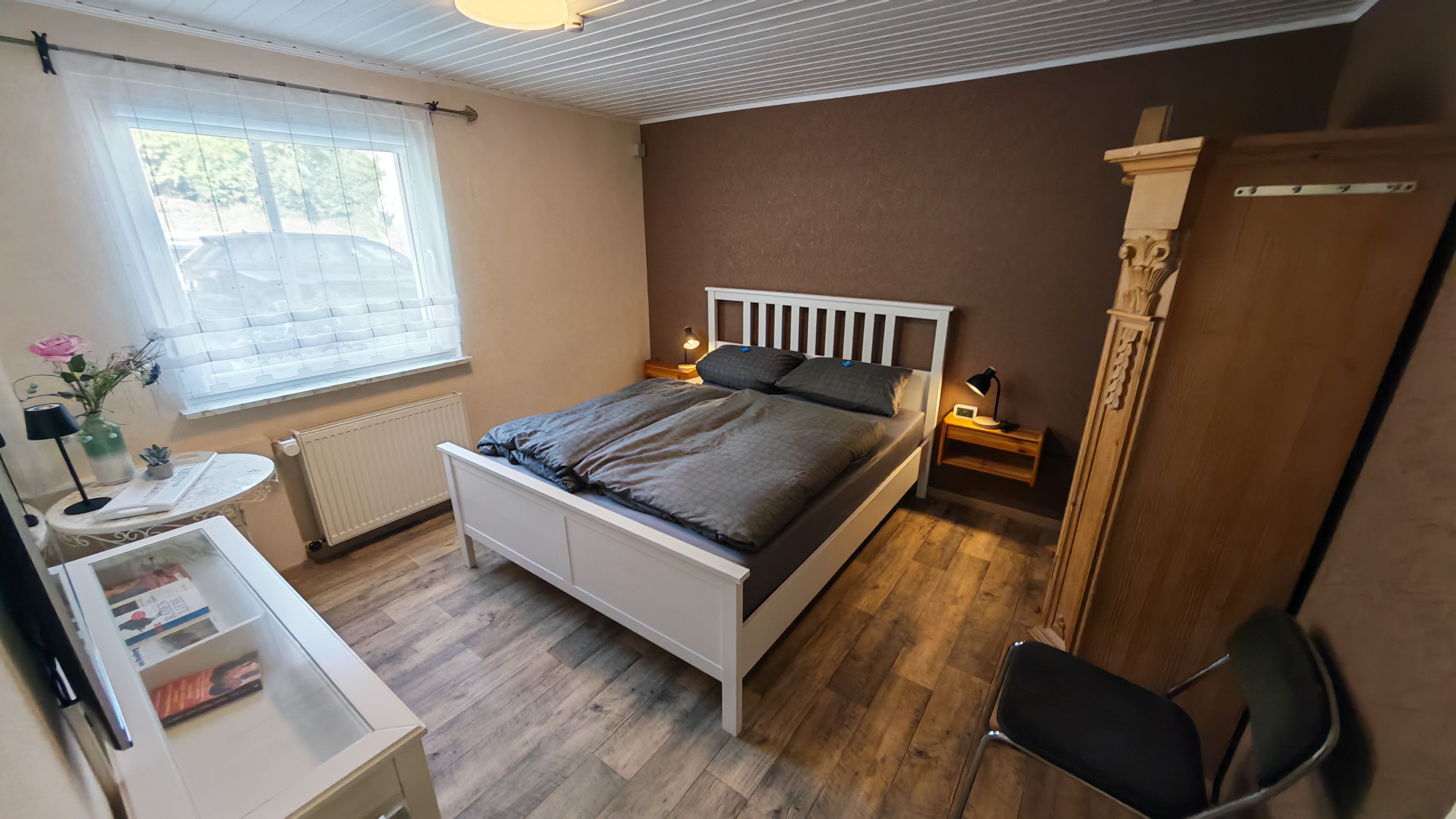 Una camera da letto con un letto, comodini, una sedia, un termosifone, una finestra con tende e un armadio in legno.