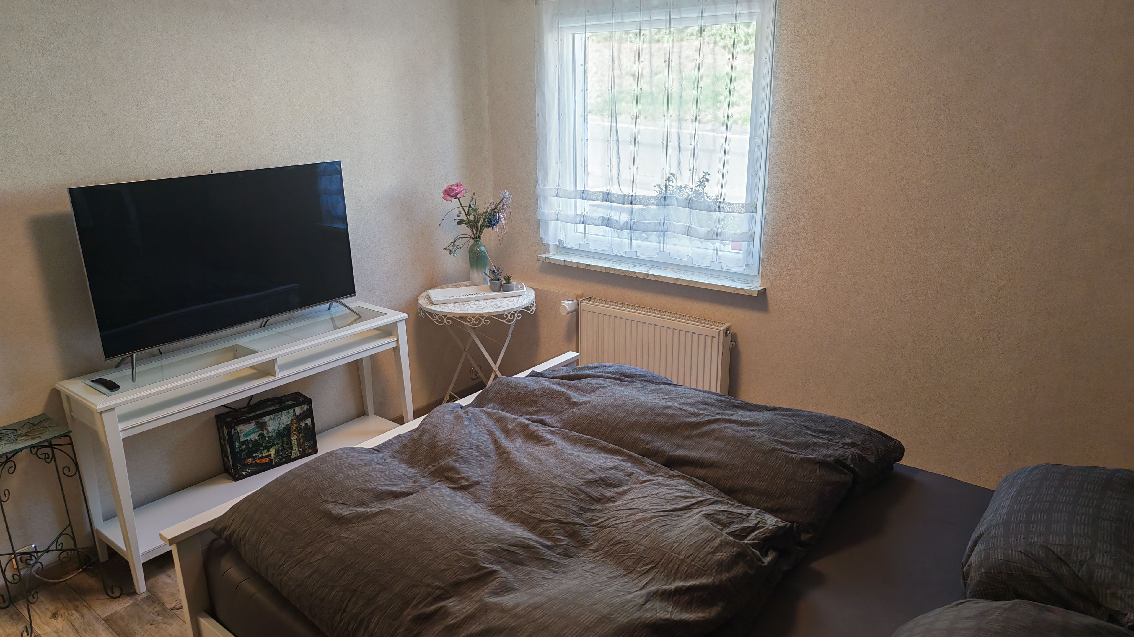Un dormitorio con cama, televisor sobre un mueble, una mesita con un florero y ventana con vista.