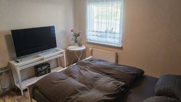 Un dormitorio con cama, televisor sobre un mueble, una mesita con un florero y ventana con vista.