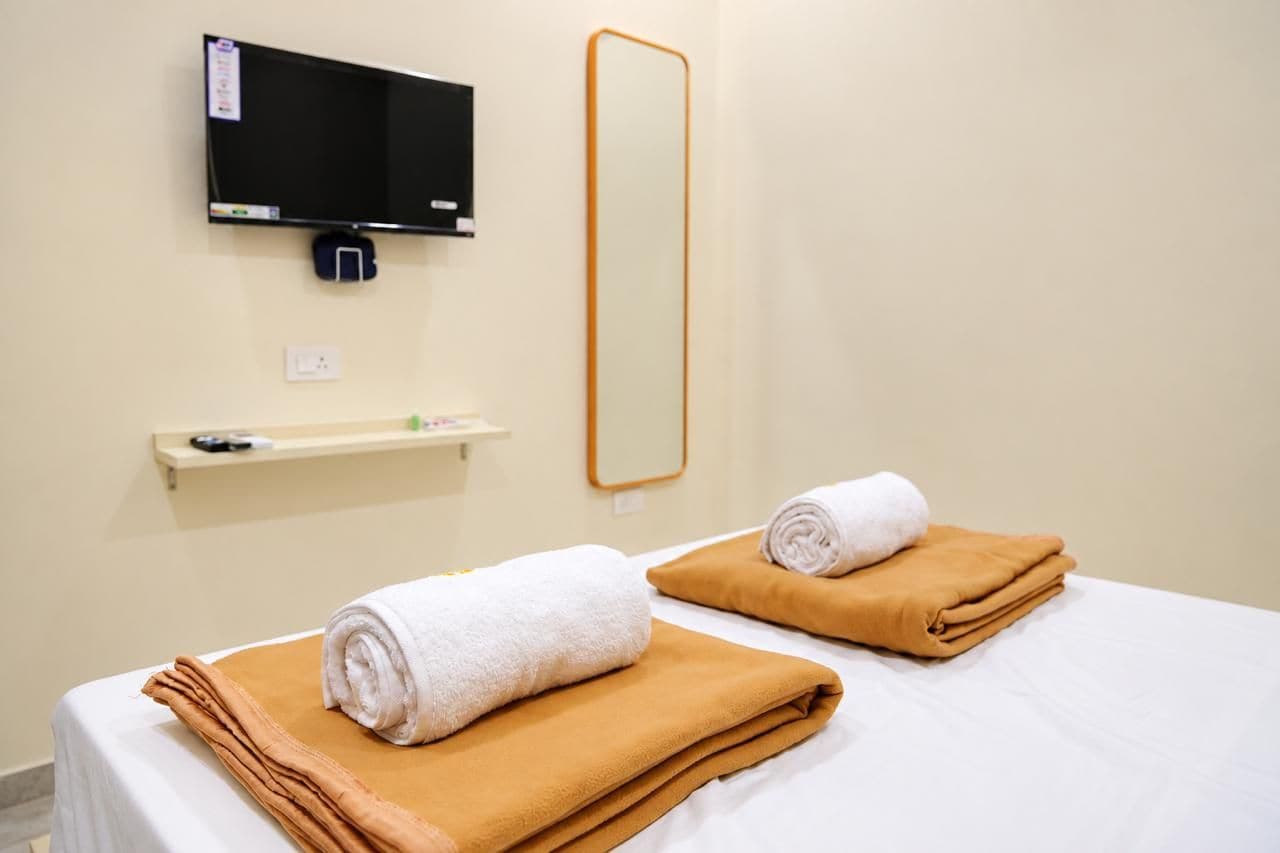 A hotel room with a bed, a TV, a mirror, and a shelf with toiletries.