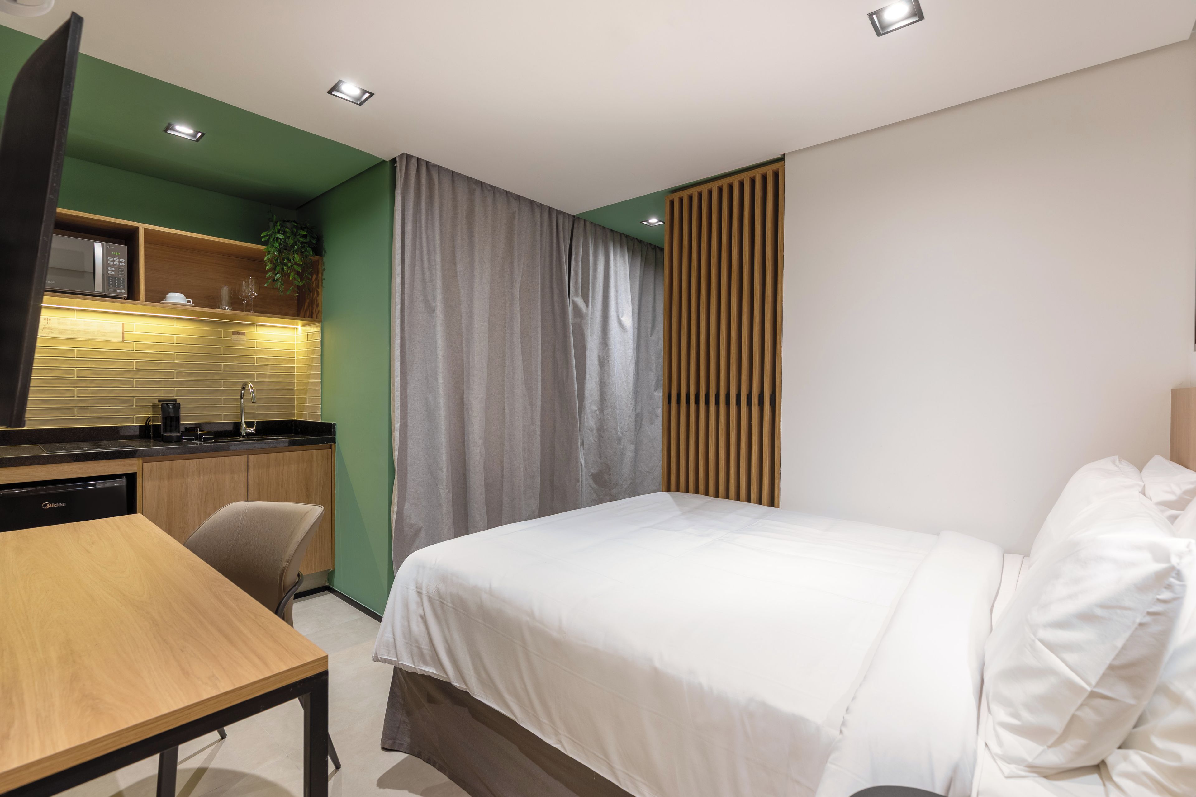 A modern hotel room with a bed, a desk, a kitchenette, and a bathroom.