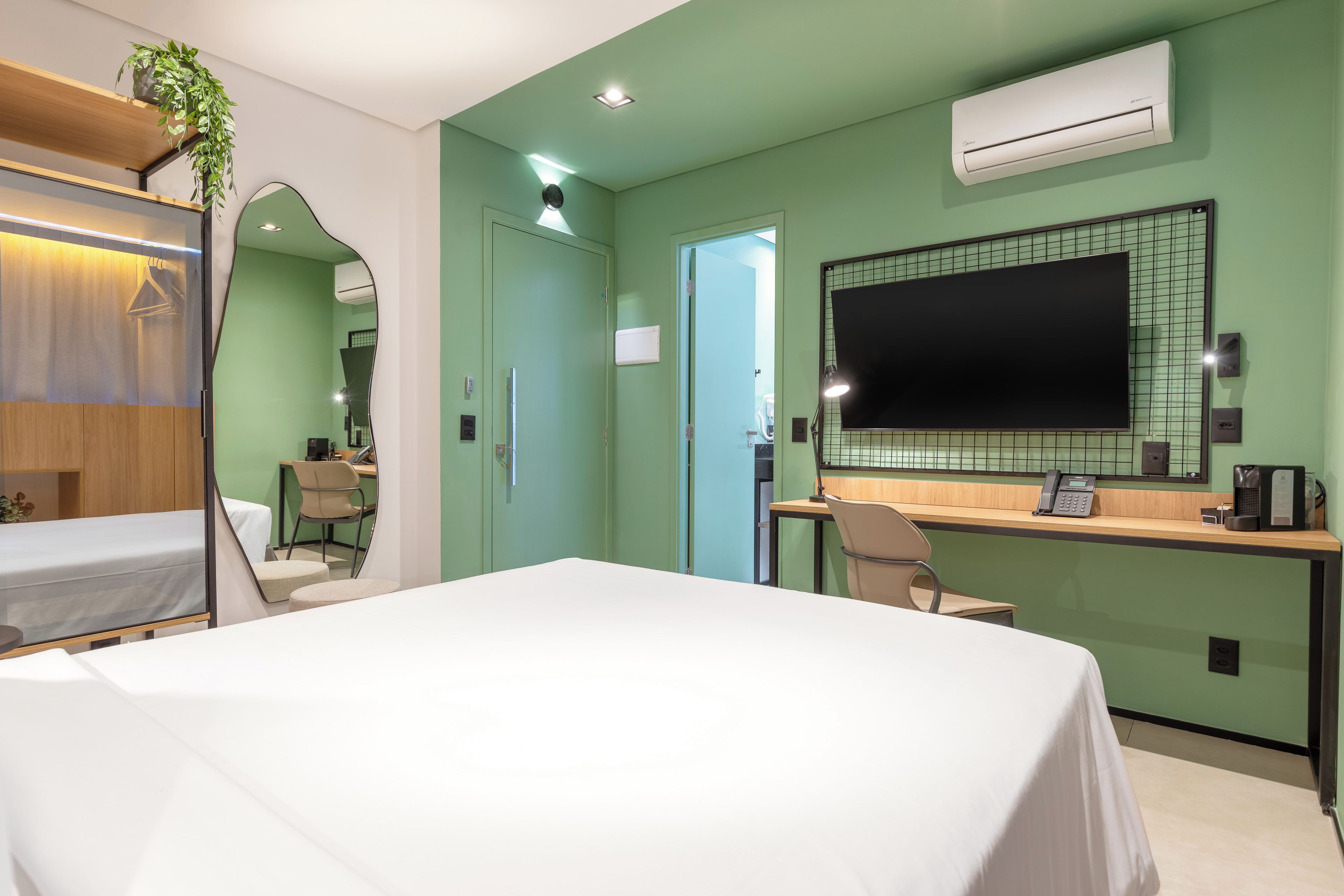 A hotel room with a bed, a desk, a chair, a mirror, and an air conditioning unit.