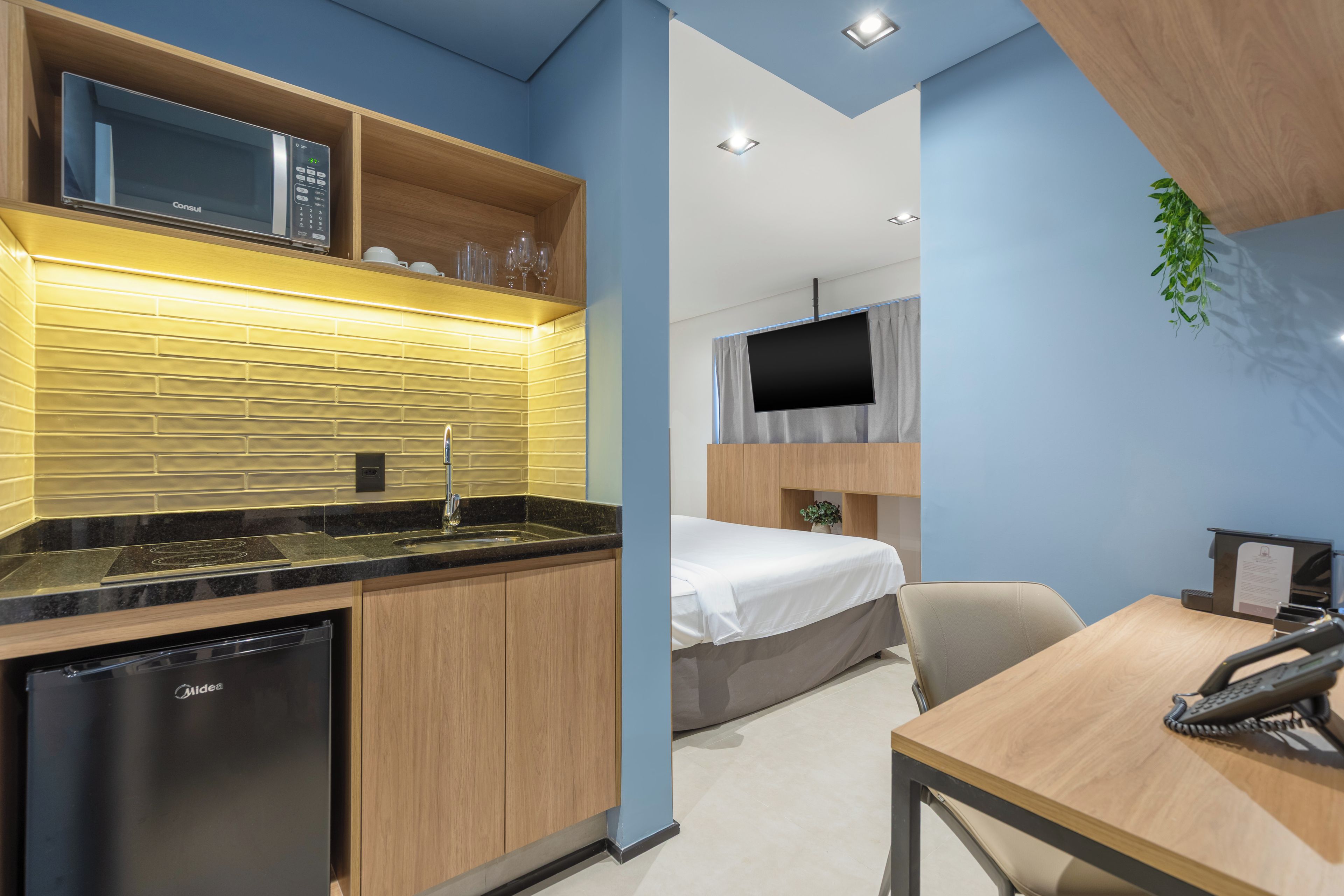 A compact hotel room with a kitchenette, a bed, a desk, and a television.