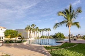A swimming pool with palm trees, a building, and a clear sky. - Casa Sirenas (Cabo San Lucas)