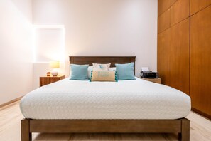 Een netjes opgemaakt bed met houten hoofdbord, een nachtkastje met een lamp en een wandplank.