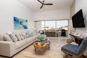 Una sala de estar moderna con sofá, mesa de centro y una silla. Se ve una ventana grande con vistas a edificios y un balcón.