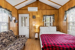 A cozy room with a bed, a sofa, a small table with a lamp, and a window with curtains. - One Room Parkview Cabin - Sleeps 2 to 4 guests. (Gore)