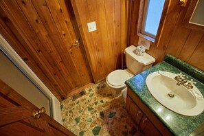 Ein Badezimmer mit einer Holztür, einer Toilette, einem Waschbecken mit grüner Arbeitsplatte und einem fußbodenähnlichen Steinmuster.