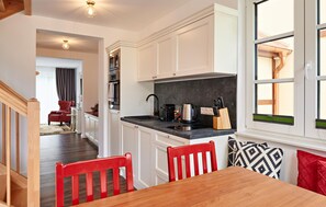 Cocina moderna con armarios blancos, encimera negra y zona de comedor con mesa de madera y sillas rojas.