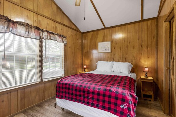 Ett rum med träpanel på väggarna, en säng, två sänglampor, ett fönster med persienner och en taklampa.