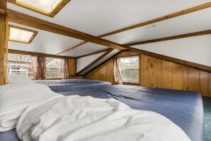 Ein Zimmer mit schrägen Wänden, Holzbalken, zwei Betten mit weißer und blauer Bettwäsche sowie Fenstern mit Vorhängen.
