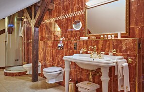 A bathroom with a marble wall, a white sink with gold fixtures, a toilet, and a shower enclosure. - 99 - Lux-App Maxi Alexandrine (Kuhlen-Wendorf)