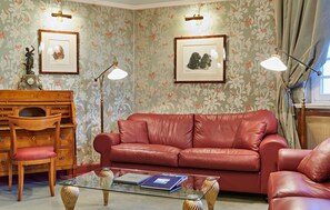 Une pièce comprenant un canapé en cuir rouge, un bureau en bois avec une chaise, une table basse en verre avec des livres et deux lampes fixées au mur.
