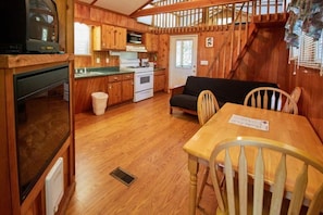 A compact kitchen with wooden cabinets, a stove, and a sink. There is a dining table with chairs and a sofa in the living area. - Two Room Trout Pond Cabin - Sleeps 6 to 8 guests, ADA Accessible (Gore)