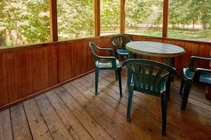 A covered outdoor area with a wooden floor, a round table, and green plastic chairs. - Two Room Trout Pond Cabin - Sleeps 6 to 8 guests, ADA Accessible (Gore)