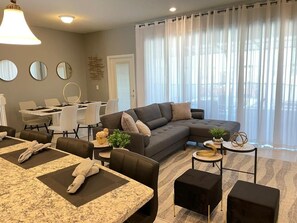 Ein modernes Wohnzimmer mit Ecksofa, Esstisch und Stühlen. Ein kleiner runder Tisch mit Pflanze und Dekorationselementen steht dort. Das Zimmer verfügt über große Fenster mit durchsichtigen Vorhängen.