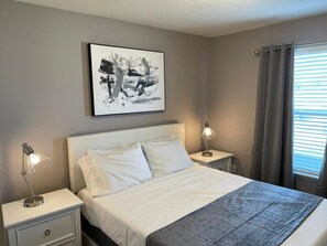 Ein Schlafzimmer mit einem Bett, zwei Nachttischen mit Lampen, einem Wandbild und einem Fenster mit Vorhängen.