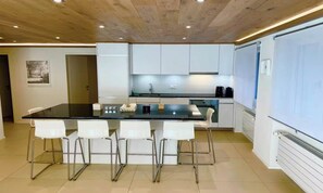 A modern kitchen with a central island, white bar stools, and a large window. - COMBINING CONTEMPORARY DESIGN WITH BREATHTAKING VIEWS OF THE ICONIC MATTERHORN (Zermatt)