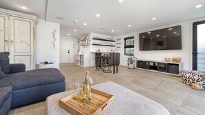 A modern living room with a sofa, a dining area with bar stools, a kitchen with a wine rack, and a large TV mounted on the wall. - Saltaire by Avantstay Rooftop Deck + Firepit + Wet Bar Walk to Beach + Trail (Marina del Rey)