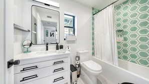 Ein Badezimmer mit weißem Waschbecken, Spiegel und Toilette, einer Duschkabine mit geometrischem Muster sowie einem Fenster mit der Aufschrift 'BATH'.